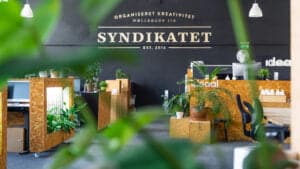 Modern coworking space at Syndikatet, featuring wooden desks, greenery, and a stylish black wall with the company name and establishment date. Ideal for fostering creativity and collaboration.