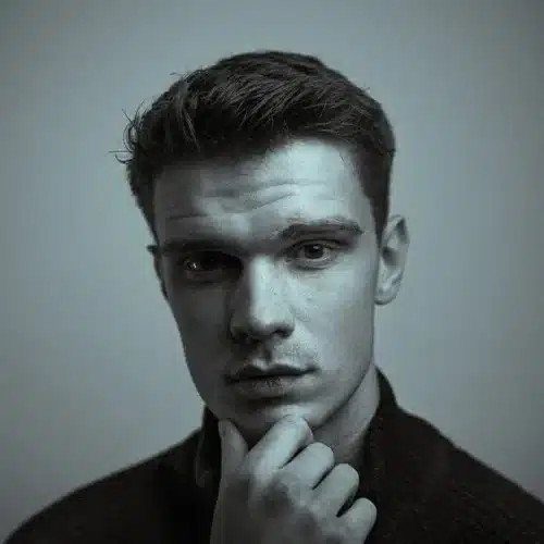Portrait of a young man with short, textured hair, thoughtfully resting his chin on his hand. The image features a moody, monochromatic color scheme, highlighting his serious expression and strong facial features against a soft background.