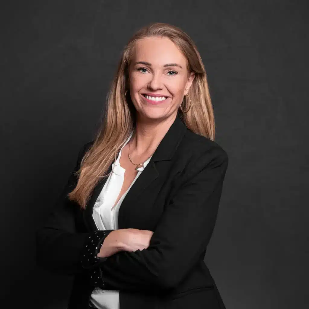 Professional woman with long hair smiling confidently in a black blazer and white blouse, standing against a dark background. Ideal for business and corporate contexts.