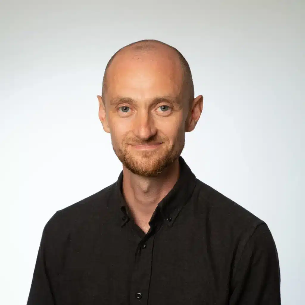Portrait of a man with a bald head and short beard, smiling against a light background, wearing a black shirt.