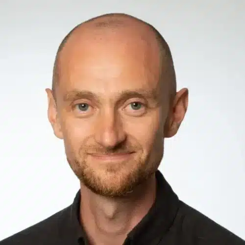 Portrait of a man with a bald head and short beard, smiling against a light background, wearing a black shirt.