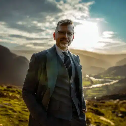En velklædt mand i jakkesæt står selvsikkert i et naturskønt landskab med bjerge og daler i baggrunden. Solen skinner blidt og skaber en varm atmosfære. Manden har briller og et venligt smil, hvilket giver en følelse af selvsikkerhed og ro i den åbne, naturskønne ramme.