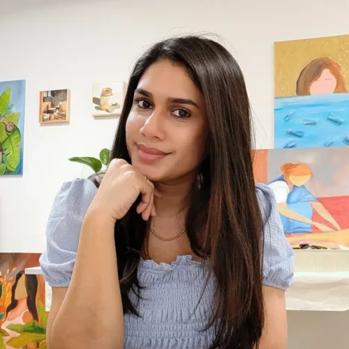 En ung kvinde sidder i et kunstnerisk atelier med en eftertænksom mine. Hun bærer en bluse med rynker og har langt, mørkt hår. Bag hende hænger farverige malerier, der viser natur og menneskelige figurer. Rummet er lyst og inspirerende, hvilket skaber en kreativ atmosfære.