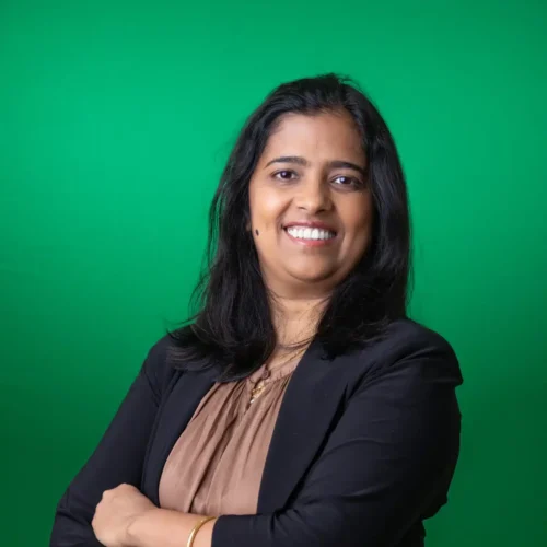 En kvinde med langt, sort hår står foran en grøn baggrund, iført en sort jakke og en brun bluse. Hun smiler og har armene korslagt, hvilket udstråler selvtillid og venlighed. Billedet har en positiv og professionel stemning, der appellerer til et bredt publikum.