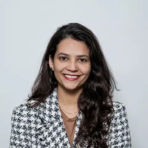 En kvinde med langt, mørkt hår smiler venligt mod kameraet. Hun bærer en sort-hvid ternet jakke og en enkel bluse. Baggrunden er lys og neutral, hvilket fremhæver hendes ansigt og udstråling. Stemningen er professionel og imødekommende, hvilket skaber en positiv atmosfære.