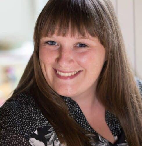 Portræt af Line Elmegaard-Simonsen. En kvinde med langt, brunt hår og pandepony, der smiler venligt og direkte ind i kameraet. Hendes hår er glat og falder naturligt ned over skuldrene. Hun har en lys hudtone og bærer en sort bluse med hvide prikker og et blomstermønster i hvidt og gråt. Baggrunden er ufokuseret, men det ser ud til at være et rum med lys farvet væg og muligvis et vindue, der giver en blød belysning. Udtrykket er varmt og imødekommende.