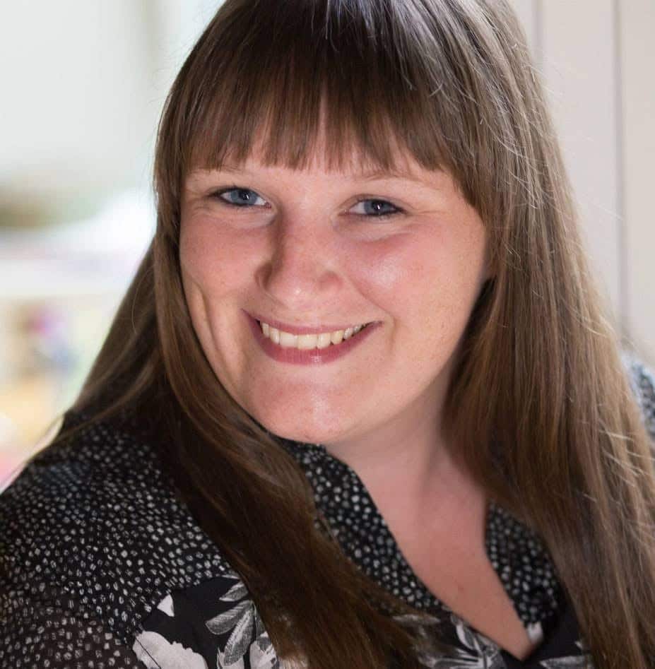 Portræt af Line Elmegaard-Simonsen. En kvinde med langt, brunt hår og pandepony, der smiler venligt og direkte ind i kameraet. Hendes hår er glat og falder naturligt ned over skuldrene. Hun har en lys hudtone og bærer en sort bluse med hvide prikker og et blomstermønster i hvidt og gråt. Baggrunden er ufokuseret, men det ser ud til at være et rum med lys farvet væg og muligvis et vindue, der giver en blød belysning. Udtrykket er varmt og imødekommende.